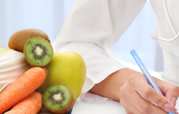 picture of hand in white lab coat holding pen with some kiwi and carrots